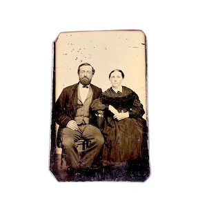 May include: A vintage sepia-toned photograph of a seated couple. The man wears a dark suit, vest, and bow tie, with a full beard. The woman is in a dark dress with a lace collar. The image has a slightly aged appearance.