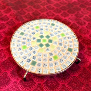 Puede incluir: Salvamanteles redondo de mosaico con marco de metal plateado y patas cortas. El diseño del mosaico presenta pequeñas baldosas cuadradas en tonos verdes, azules y crema. El salvamanteles protege las superficies de objetos calientes.