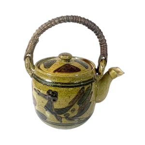 May include: A ceramic teapot with a woven handle and a lid. The teapot is primarily olive green with dark brown and black painted designs. The spout is gold-colored.