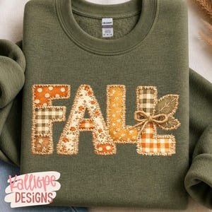 May include: An olive green crewneck sweatshirt featuring the word FALL in large, patchwork-style letters. The letters are composed of various fabric patterns including orange polka dots, brown gingham, and floral prints, finished with a jute twine bow.