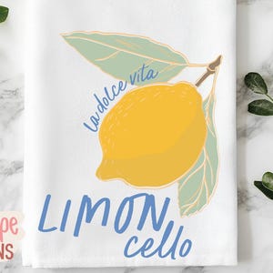 K&ouml;nnte beinhalten: Wei&szlig;es K&uuml;chentuch mit einer gelben Zitronenillustration und den Worten "La dolce vita" und "Limoncello" in Blau. Die Zitrone hat gr&uuml;ne Bl&auml;tter und einen braunen Stiel. Das Tuch liegt auf einer Marmoroberfl&auml;che.