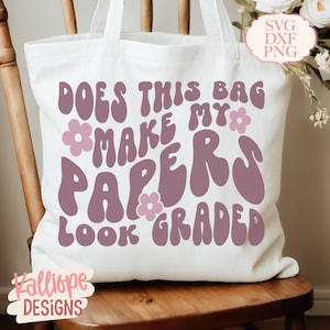 Może przedstawiać: Biała torba typu tote z napisem "DOES THIS BAG MAKE MY PAPERS LOOK GRADED" w kolorze fioletowym, z kwiatowymi akcentami. Torba stoi na drewnianym krześle. Torba ma uchwyt.