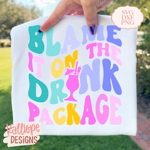 May include: White t-shirt with the phrase "Blame It On The Drink Package" in colorful, retro-style lettering. The design includes a cocktail illustration. The words "SVG DXF PNG" and "Kalliope Designs" are also visible.