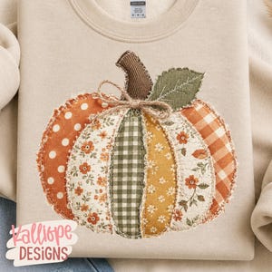 May include: A beige crewneck sweatshirt featuring a large, central pumpkin applique made of various fabric patches in orange, green, and white with polka dot, floral, and gingham patterns, topped with a brown corduroy stem and a jute twine bow.