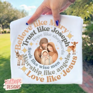 May include: White t-shirt featuring a circular design with the text "Believe like Mary, Trust like Joseph, Love like Jesus." The design includes illustrations of the Holy Family, a donkey, and a calf. The brand name "Falliope Designs" is at the bottom.