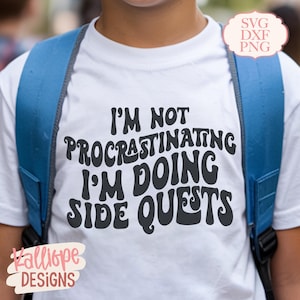 Könnte beinhalten: Weißes T-Shirt mit dem schwarzen Text "I'M NOT PROCRASTINATING I'M DOING SIDE QUESTS" in einer Retro-Schriftart. Das T-Shirt wird mit einem blauen Rucksack getragen. Das Bild enthält auch den Text "SVG DXF PNG" und "Kalliope Designs".