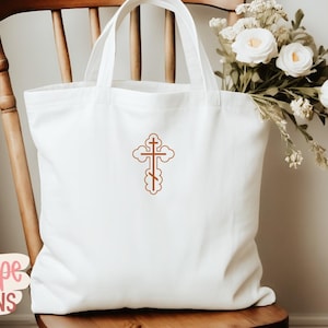 Op de afbeelding: Een witte draagtas met een bruin geborduurd kruisontwerp. De tas staat op een houten stoel, met een boeket witte bloemen op de achtergrond. De tas heeft een eenvoudig ontwerp, perfect voor het dragen van spullen.
