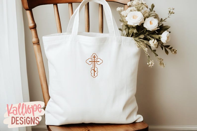 Op de afbeelding: Een witte draagtas met een bruin geborduurd kruisontwerp. De tas staat op een houten stoel, met een boeket witte bloemen op de achtergrond. De tas heeft een eenvoudig ontwerp, perfect voor het dragen van spullen.