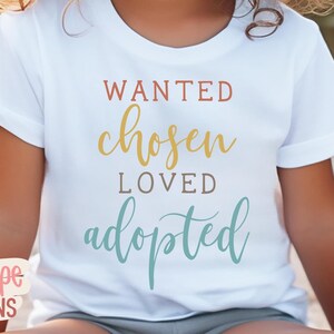 May include: White t-shirt with the words "WANTED, chosen, LOVED, adopted" in various colours. The text is in a handwritten style font. The shirt is short-sleeved.