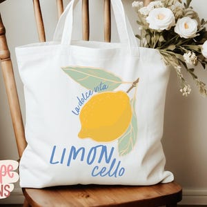K&ouml;nnte beinhalten: Wei&szlig;er Stoffbeutel mit einer Zitronenillustration und den Worten "La dolce vita" und "Limoncello" in Blau. Die Tasche steht auf einem Holzstuhl mit einem Strau&szlig; wei&szlig;er Blumen.