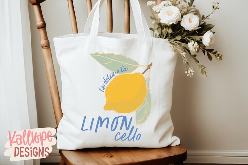 K&ouml;nnte beinhalten: Wei&szlig;er Stoffbeutel mit einer Zitronenillustration und den Worten "La dolce vita" und "Limoncello" in Blau. Die Tasche steht auf einem Holzstuhl mit einem Strau&szlig; wei&szlig;er Blumen.