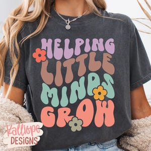 May include: Dark gray t-shirt with the text "Helping Little Minds Grow" in colorful, retro-style lettering. The design includes flower accents. The shirt is a casual wear item.