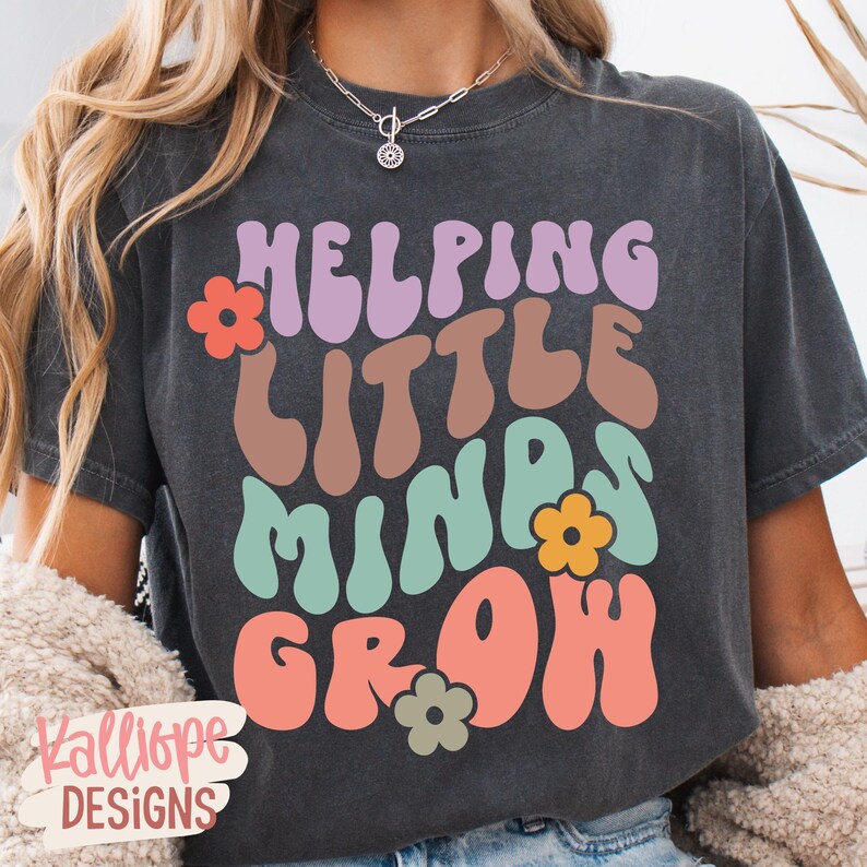 May include: Dark gray t-shirt with the text "Helping Little Minds Grow" in colorful, retro-style lettering. The design includes flower accents. The shirt is a casual wear item.