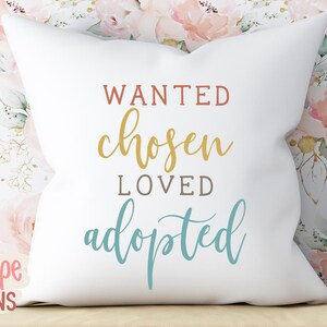 May include: A white square cushion with the words "WANTED, chosen, LOVED, adopted" in various colours. The cushion is on a wooden surface with a floral background and a stuffed toy.