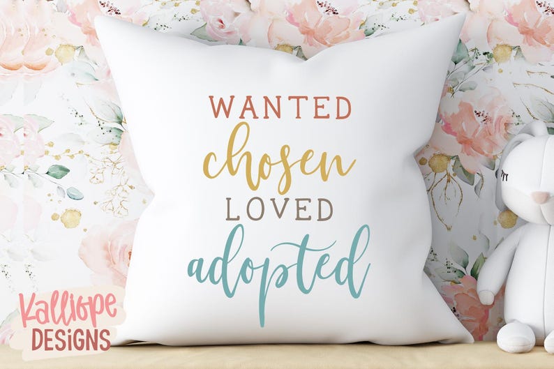 May include: A white square cushion with the words "WANTED, chosen, LOVED, adopted" in various colours. The cushion is on a wooden surface with a floral background and a stuffed toy.