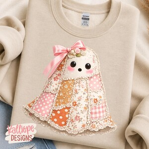 May include: A beige crewneck sweatshirt featuring a graphic of a ghost made of patchwork fabric in shades of pink, orange, and white. The ghost has large black eyes, pink cheeks, and a pink satin bow on its head. The text Kalliope DESIGNS is visible.