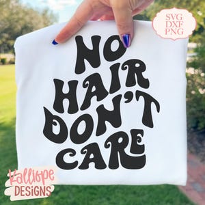 May include: A white t-shirt with the phrase "NO HAIR DON'T CARE" in large, bold, black letters. The design has a retro, groovy font style. The shirt is held up by a hand with blue nail polish. The bottom left corner has the text "Kalliope DESIGNS".