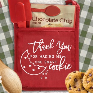 Pode incluir: Uma luva de forno vermelha com um bolso contendo um pacote de mistura para biscoitos com pepitas de chocolate, uma espátula vermelha e uma colher vermelha. A luva tem o texto "Thank You For Making Me One Smart Cookie" e um gráfico de biscoito. Biscoitos com pepitas de chocolate em primeiro plano.