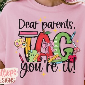 May include: Pink t-shirt with the text "Dear parents, TAG you're it!" in black script. The design includes a red apple, green ruler, and school supplies. The shirt has a crew neck and short sleeves.