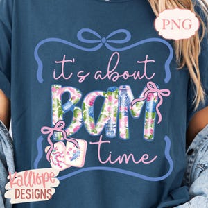 May include: A navy blue t-shirt with the phrase "it's about BAM time" in pink script. The word "BAM" is filled with a floral pattern of pink and blue flowers. A pink bow and two cards with floral designs are also featured.