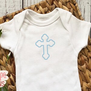 Op de afbeelding: Witte babybody met een lichtblauw kruisontwerp. De body ligt op een gevlochten mand. De tekst "Kalliope Designs" is linksonder zichtbaar. Er zijn ook een paar takjes witte bloemen.