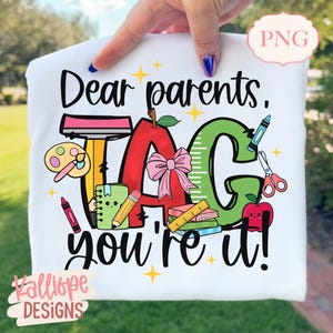 May include: White t-shirt with a colorful school-themed graphic. The design includes an apple, the word "TAG", a ruler, a crayon, and the text "Dear parents, you're it!" in black script. The design also includes a pink bow.