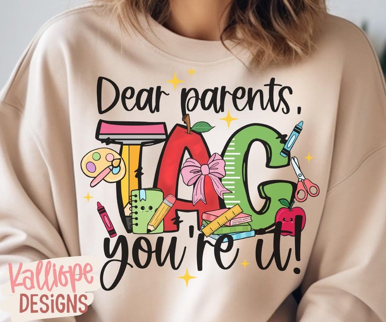 May include: Beige sweatshirt with a colorful graphic that reads "Dear parents, TAG, you're it!" The design includes an apple, school supplies, and stars. The brand name "Kalliope DESIGNS" is at the bottom.