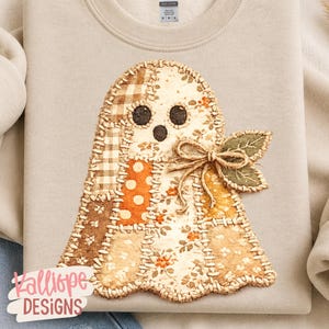 May include: A beige sweatshirt featuring a central ghost-shaped applique made of various fabric patches in brown, orange, and cream tones. The ghost has three dark circular eyes and mouth, a jute twine bow, and two green fabric leaves attached.