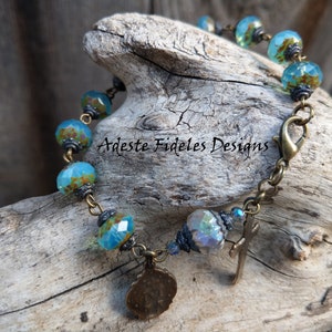 May include: A bracelet featuring faceted turquoise and brown glass beads, antique bronze-toned metal spacers, a lobster claw clasp, and two hanging charms, one circular and one cross-shaped. The text Adeste Fideles Designs is visible in the center.