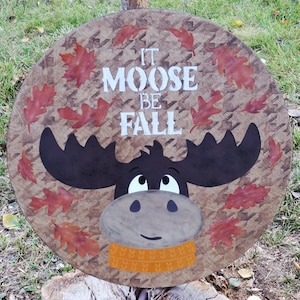 May include: Round wooden sign with a moose illustration and the text "It Moose Be Fall." The sign features a brown background with a houndstooth pattern, a cartoon moose with a yellow scarf, and red autumn leaves.