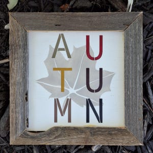 Könnte beinhalten: Quadratisches Holzschild mit dem Wort "HERBST" in verschiedenen Farben. Die Buchstaben sind vertikal über einem verblassten Blattmuster angeordnet. Der Rahmen besteht aus verwittertem Holz.