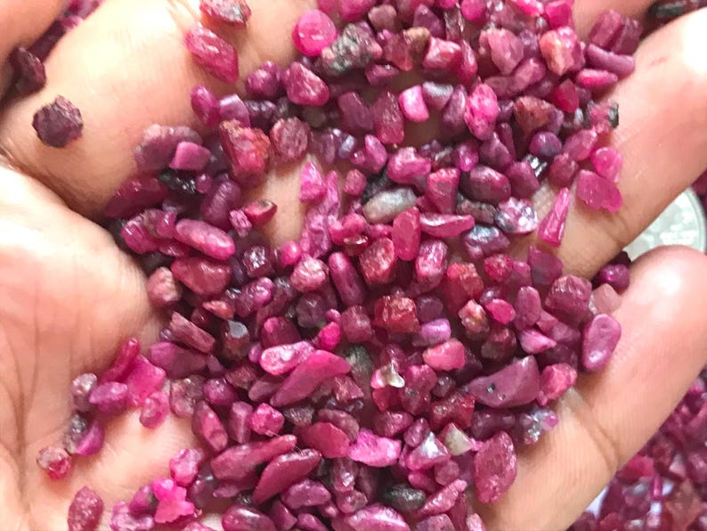 Op de afbeelding: Een verzameling kleine, ruwe, ongepolijste robijn-edelstenen in verschillende tinten diep roze en levendig rood. De stenen zijn onregelmatig van vorm en grootte en hebben een natuurlijk, getextureerd oppervlak met doorschijnende delen.