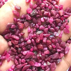 Op de afbeelding: Een verzameling kleine, ruwe, ongepolijste robijn-edelstenen in verschillende tinten dieproze en levendig rood. De stenen hebben onregelmatige, grillige vormen en een natuurlijke, matte tot licht glanzende textuur in de palm van een hand.