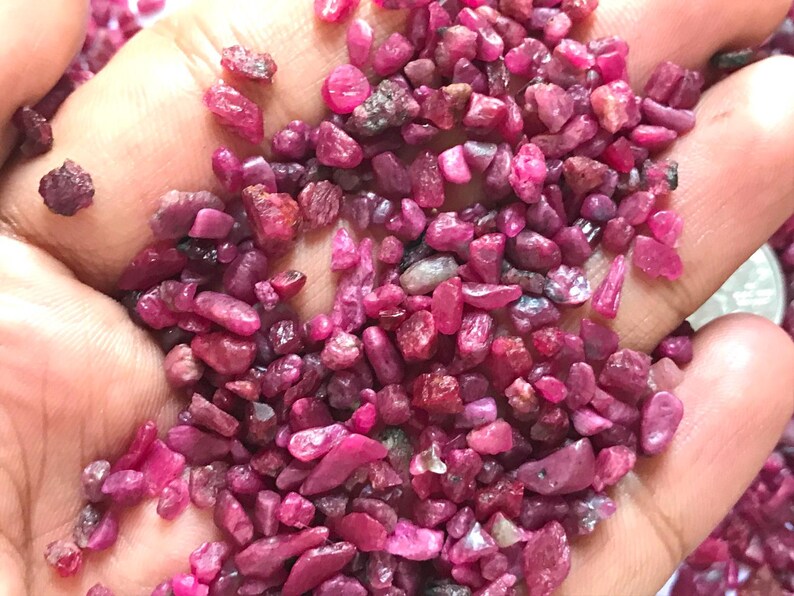 Op de afbeelding: Een verzameling kleine, ruwe, ongepolijste robijn-edelstenen in verschillende tinten dieproze en levendig rood. De stenen hebben onregelmatige, grillige vormen en een natuurlijke, matte tot licht glanzende textuur in de palm van een hand.