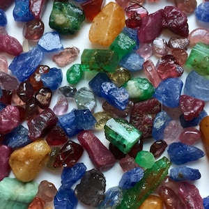 Peut inclure: Une collection de pierres précieuses brutes et non taillées de formes et de tailles variées, présentant un mélange vibrant de couleurs incluant le rouge profond, le bleu vif, le vert émeraude et le jaune doré, éparpillées sur une surface blanche.