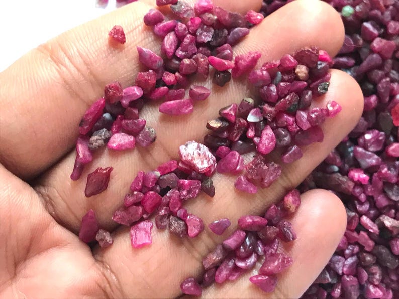 Op de afbeelding: Een verzameling kleine, ruwe, getrommelde robijn-edelstenen in verschillende tinten diep roze en levendig rood. Deze natuurlijke, ongepolijste mineraalfragmenten hebben onregelmatige vormen en texturen en zien er doorschijnend tot ondoorzichtig uit.