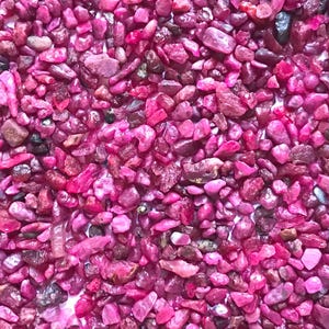 Op de afbeelding: Een verzameling kleine, onregelmatige, getrommelde edelsteenfragmenten in levendige tinten magenta en dieproze. De stenen hebben een gepolijste, glanzende textuur met vari&euml;rende niveaus van doorschijnendheid en af en toe donkere insluitingen.