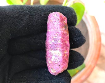 May include: A raw, elongated ruby crystal with a vibrant magenta-pink hue and a textured, uneven surface. The gemstone displays natural crystalline facets and small mineral inclusions, showcasing a deep, saturated color throughout its structure.