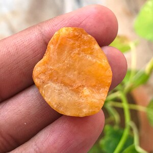 以下が含まれることがあります： 不規則な形の半透明のオレンジ色の宝石。表面は粗く質感があり、内部の結晶構造が見えます。琥珀色と金色の色合いが混ざり合い、輪郭はわずかに不均一で丸みを帯びています。