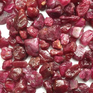 May include: A selection of raw ruby gemstones in various sizes and shapes. The stones exhibit a spectrum of deep red colours, with some having a polished, reflective surface and others appearing rough and textured. The gemstones are scattered across the image.