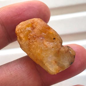 May include: A raw, unpolished gemstone with a translucent, golden-orange hue. The stone features an irregular, multifaceted surface with natural textures, internal inclusions, and varying shades of amber and honey throughout its crystalline structure.