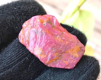May include: A raw, unpolished ruby gemstone with a vibrant pinkish-red hue and irregular surface texture. The stone features visible orange and brown mineral inclusions throughout its crystalline structure, showcasing a natural, jagged, and multifaceted shape.