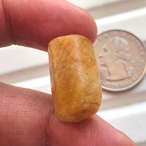 Peut inclure: Une petite perle cylindrique en pierre précieuse jaune doré translucide avec une surface naturelle texturée. La perle est tenue à la verticale, placée à côté d'une pièce de monnaie américaine pour l'échelle, soulignant sa taille compacte.