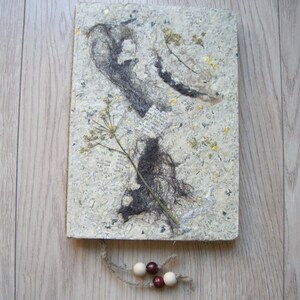 May include: A handmade journal with a textured, off-white cover adorned with pressed botanicals, dark grey accents, and gold flecks. A sprig of dried flowers and a string with wooden beads dangle from the bottom. The journal is approximately 15 x 20 cm.