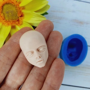 Può includere: Una piccola scultura di un viso umano, pallida e non dipinta, con gli occhi chiusi e un'espressione neutra. La scultura è realizzata in un materiale liscio e bianco ed è tenuta in una mano.
