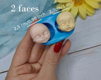 Könnte beinhalten: Eine blaue Silikonform mit zwei Vertiefungen in Form von Gesichtern. Die Form ist 2,7 cm/1 Zoll breit und 2,5 cm/0,98 Zoll hoch. Die Form wird in einer Hand gehalten.