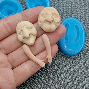 Puede incluir: Dos caras sin pintar y dos brazos sin pintar hechos de arcilla polimérica. Las caras tienen detalles elaborados, incluyendo arrugas y una nariz grande. Los brazos están doblados en el codo. Las piezas de arcilla están sentadas en una mano, y hay un molde de silicona azul en el fondo.