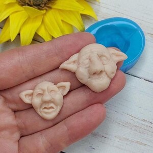 May include: Two small, beige, sculpted faces with pointed ears and distinct facial features, alongside a blue silicone mold. The faces have detailed noses, eyes, and mouths, appearing as individual castings created from the circular blue mold.
