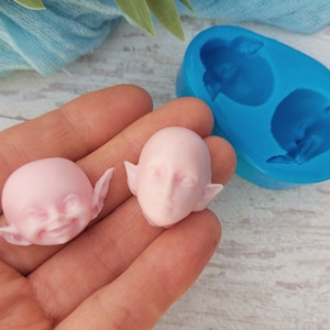 May include: Two pink silicone moulds for making fairy or elf heads. The moulds are in the shape of a head with pointy ears. The moulds are on a white wooden surface.
