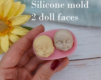 Könnte beinhalten: Eine rosa Silikonform mit zwei Abdrucken von Puppenköpfen. Die Form wird in einer Hand gehalten. Der Text "Silikonform 2 Puppenköpfe" ist über der Form sichtbar.
